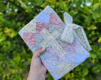 Puede incluir: Un cuadrado acolchado hecho a mano en colores pastel rosa, azul y amarillo, con un estampado floral. El edredón está adornado con un lazo de encaje color crema. El artículo se sostiene en una mano sobre un fondo verde borroso.
