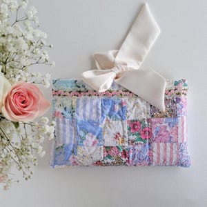 May include: A small patchwork fabric clutch with a white satin bow. The clutch is made of various floral and striped fabrics in shades of pink, blue, and white.