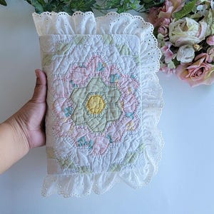 May include: A rectangular fabric book cover featuring a quilted floral pattern in pastel pink, pale green, and yellow. The cover is edged with a white eyelet lace ruffle trim, providing a decorative border around the stitched hexagonal design.