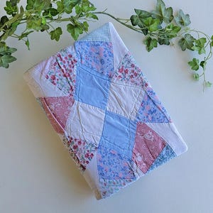 May include: A folded patchwork quilt featuring a diamond pattern composed of light blue, dusty rose, and white fabric squares. The quilt displays a mix of solid colors and floral prints, with white fabric borders separating the individual sections.