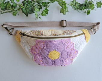 Puede incluir: Una riñonera acolchada con un diseño de patchwork de telas florales en tonos lavanda, amarillo y blanco. El bolso cuenta con un cierre de cremallera superior, una correa ajustable de color beige con herrajes metálicos y textura cosida.