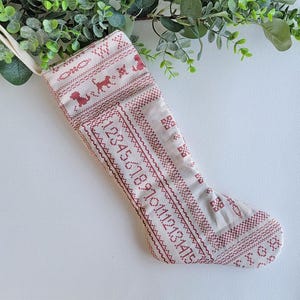 May include: A cream-colored Christmas stocking with red embroidered details. The design includes numbers, dogs, and geometric patterns. A loop is attached for hanging. The stocking is decorated with a festive theme.