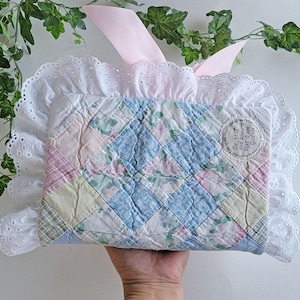 May include: A rectangular quilted fabric cover featuring a diamond-patterned patchwork of pastel blue, pink, yellow, and green floral prints. The cover is edged with a wide white eyelet lace ruffle and includes a light pink ribbon tie at the top.
