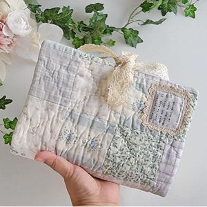 May include: A quilted fabric book cover featuring a patchwork design of light blue, green, and white floral and plaid patterns. A cream-colored lace ribbon is tied at the top. A rectangular fabric patch with the text Love is patient, love is kind.