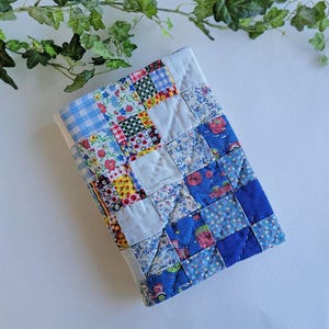 May include: A patchwork quilt in a rectangular shape, featuring a variety of fabric patterns including floral, gingham, and solid blue squares. The quilt is primarily blue, white, and red, with a quilted texture.