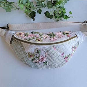 May include: A quilted fanny pack featuring a patchwork design of light gray, white, and floral fabrics. The bag is decorated with embroidered pink and green flowers and a central heart-shaped patch, finished with a beige adjustable strap.