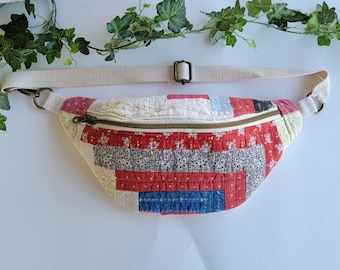 Puede incluir: Riñonera acolchada con correa y cremallera color crema. La bolsa presenta un diseño de patchwork en telas rojas, blancas, azules y rosas. La correa ajustable es color crema y mide unos 30 cm de largo.