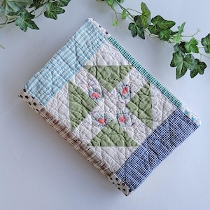 May include: A rectangular quilted fabric cover featuring a central pinwheel block pattern in sage green and white. The border consists of various fabric strips in blue gingham, brown plaid, and black and white polka dot patterns.
