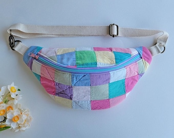 Puede incluir: Una riñonera con un diseño de parches de cuadrados de tela en tonos pastel rosa, azul, verde, amarillo y morado. El bolso cuenta con una cremallera azul claro y una correa tejida ajustable en color blanco roto con herrajes de metal oscuro.
