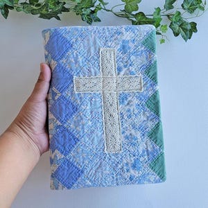 May include: A rectangular book cover featuring a quilted patchwork design in various shades of blue and white floral patterns. A cream-colored lace cross is centered on the front. The right edge is finished with a solid green fabric border.