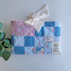 May include: A patchwork quilted clutch in shades of blue and pink, adorned with a cream-colored bow. The design features floral patterns and solid squares. A small tag reads "God is in control, His timing is perfect."