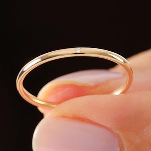 May include: A thin, polished gold band ring featuring a single, small, round, clear gemstone set flush into the top center of the metal. The ring has a smooth, rounded profile and a warm, metallic yellow gold finish that reflects light.
