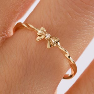 May include: A gold-colored ring with a bow design. The ring features a twisted band and a small bow with a single, clear gemstone in the center. The ring is shown on a finger.
