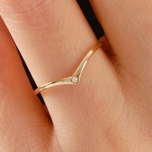 May include: A delicate gold ring with a V-shaped design. The ring features a small, clear gemstone at the center of the V. The ring is worn on a finger, showcasing its elegant and minimalist style. The ring is a piece of jewellery.