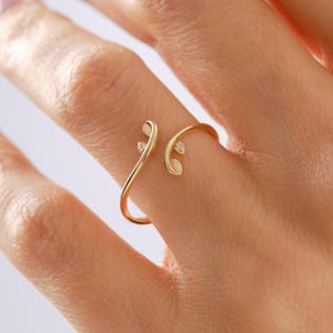 May include: A gold open-style ring featuring two delicate leaf-shaped ends, each accented with a single small, clear, teardrop-shaped gemstone. The band is thin and polished, designed to wrap partially around the finger with an open gap.