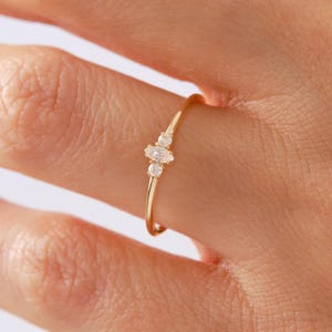 Peut inclure: Une fine bague en or ornée d'une pierre précieuse transparente centrale de taille marquise, encadrée par deux pierres rondes plus petites. L'anneau est lisse et poli, présentant un design délicat avec trois pierres alignées horizontalement.