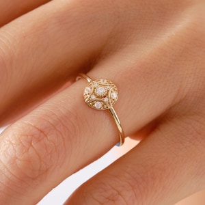 May include: A gold ring featuring a circular centerpiece with a central round gemstone surrounded by smaller accent stones, set within a textured metal frame. The band is thin and polished, showcasing a delicate design with intricate metalwork.