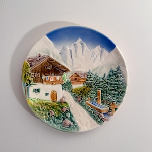 May include: A decorative ceramic plate featuring a mountain village scene. The plate depicts a chalet with a brown roof, a road, and a water trough, all set against a backdrop of snow-capped mountains and a blue sky.