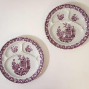 May include: Two white ceramic divided plates featuring a purple transferware pattern. Each plate has three sections and a wide border decorated with floral motifs. The center of each plate displays a detailed landscape scene with a fountain.