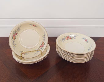 May include: A set of cream-colored vintage bowls with floral patterns in pink, blue, and yellow. The bowls have a gold rim and are stacked on a dark wooden surface. Some bowls are displayed on a gold-colored metal stand.