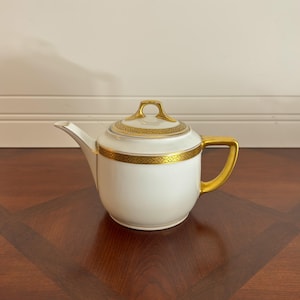 May include: A white porcelain teapot featuring a gold handle, a gold spout tip, and a gold lid finial. The body and lid are decorated with a wide, intricate gold band featuring a repeating geometric pattern around the upper rim.