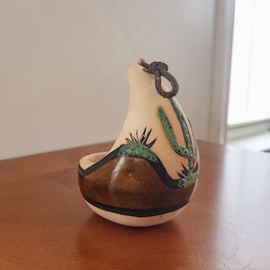 May include: A hand-painted, gourd-shaped decorative item with a desert landscape design. The item features a cream-colored body with brown, black, and green painted details, including cacti and a mountain scene. A brown rope loop is attached at the top.