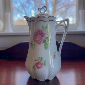 May include: White porcelain coffee pot with a lid, featuring pink rose floral designs and gold trim. The pot has a slender, elegant shape with a handle and a decorative finial on the lid. A classic piece for serving coffee or tea.