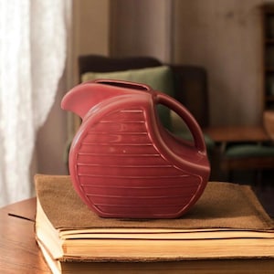 May include: A red ceramic pitcher with a ribbed design. The pitcher is sitting on a stack of books.