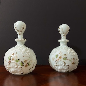 May include: Two antique white glass perfume bottles with ornate floral and relief designs. Each bottle features a bulbous body, a decorative stopper, and accents of gold and green. The bottles are displayed on a dark wooden surface.