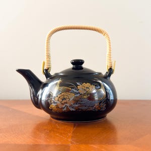 May include: Black ceramic teapot with a woven handle and a lid. The teapot is decorated with gold and silver floral and bird designs. The handle is made of a light tan woven material.