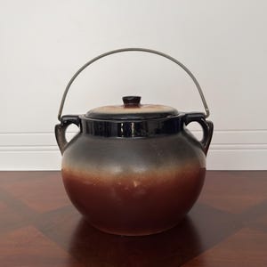 May include: A ceramic pot with a lid and a metal handle. The pot is a gradient of colors, transitioning from black at the top to brown at the bottom. The lid has a small knob and the handle arches over the top.