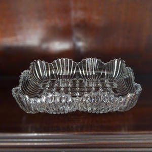 May include: A clear, rectangular glass dish with a scalloped rim. The dish has a diamond-patterned base and vertical lines along the sides. The glass is transparent, allowing light to pass through, and is set against a dark brown background.