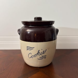 May include: A vintage cookie jar with a dark brown lid and upper section, and a cream-colored lower section. The word "Cookie" is written in cursive with blue ink. The jar has two small handles.