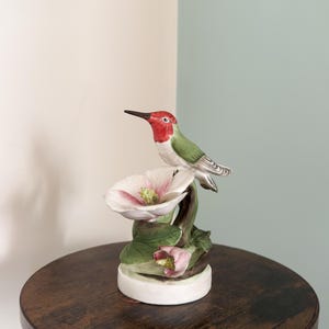May include: A porcelain figurine featuring a hummingbird with a bright red head and green wings perched on a branch above a large white flower with pink accents and green leaves, all set upon a circular white base.