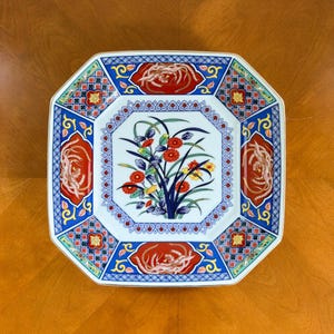 May include: An octagonal ceramic plate with a floral design. The plate features a central bouquet of red, yellow, and blue flowers. The border has red, blue, and gold accents with a floral and geometric pattern. The plate is on a wooden surface.