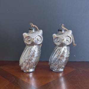 May include: Two silver-colored owl ornaments with textured surfaces. Each owl has large, round eyes and a small loop for hanging. The ornaments are made of glass and have a vintage, antique appearance. They are suitable for holiday or home decor.