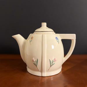 May include: Cream-colored ceramic teapot with a lid, spout, and handle. The teapot is decorated with small, colorful floral designs. The handle is rectangular, and the lid has a ribbed design. The teapot is sitting on a wooden surface.