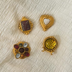 May include: Four decorative gold-tone buttons with distinct designs. One features a rectangular maroon stone, another a central heart, a third has a cluster of multi-colored cabochons, and the fourth displays a textured yellow circular inlay.
