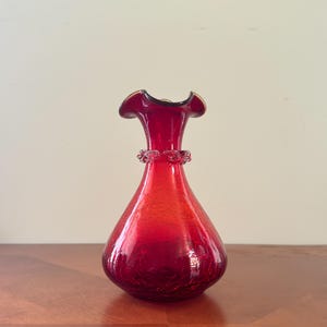 May include: A red glass vase with a ruffled rim and a decorative band around the neck. The vase has a bulbous body and a crackled texture. The vase is approximately 8 inches tall.