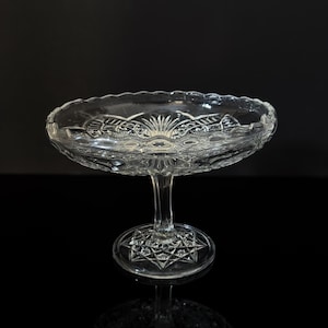 May include: A clear glass pedestal serving dish with a scalloped edge. The dish has a detailed pattern around the rim and on the base. The stem is slender, and the base is a starburst design. The dish is likely used for serving desserts or displaying items.