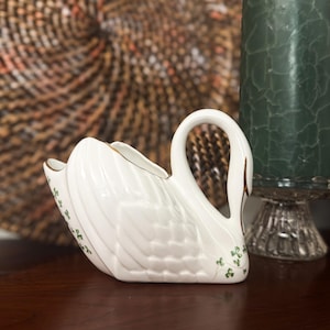 May include: White ceramic swan-shaped decorative dish with gold trim and green shamrock accents. The swan has a curved neck and is sitting on a dark wooden surface. A green candle and glass candle holder are in the background.