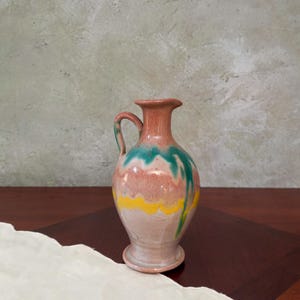 May include: A ceramic pitcher with a handle, featuring a base and a narrow neck. The pitcher is painted with a blend of colors, including pink, green, yellow, and brown. The design is a mix of dripped and painted patterns, creating a unique, handcrafted look.