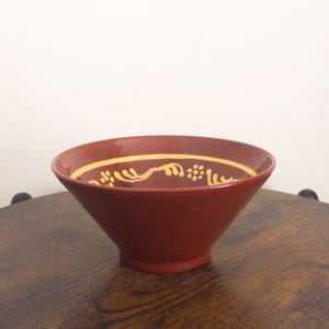 May include: A maroon-colored ceramic bowl with a flared shape. The rim features a yellow decorative band with a repeating pattern of stylized floral or leaf-like designs. The bowl sits on a wooden surface.