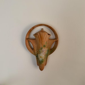 May include: A peach-colored wall pocket vase with a circular frame. The vase features a floral design in shades of white, pink, and green. The wall pocket is made of ceramic or a similar material and is designed to hold flowers or other decorative items.