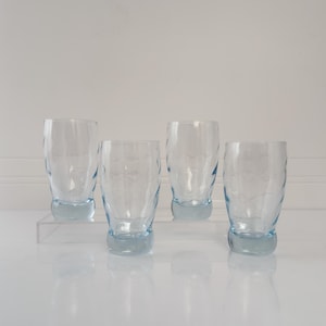 May include: Four clear, textured drinking glasses with a light blue base. The glasses have a unique wave-like pattern and are displayed on a clear surface. The glassware is ideal for serving beverages.
