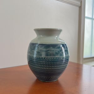 May include: A small ceramic vase with a rounded body and a flared rim. The upper section is a smooth light cream color, while the lower portion features a deep blue glaze with intricate white wavy line patterns and a textured dotted design.