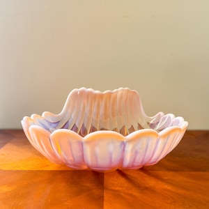 May include: A decorative, iridescent glass bowl with a scalloped rim and a central, raised design. The bowl has a soft, peachy-pink color with hints of purple and yellow. The bowl is likely used for serving or display.