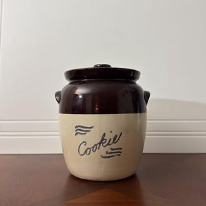 May include: A vintage brown and cream-colored cookie jar with a lid. The jar has a dark brown top and lid, with a cream-colored base. The word "Cookie" is written in cursive in dark blue on the front.