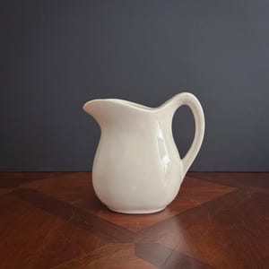 May include: Cream-colored ceramic pitcher with a rounded body and a curved handle. The pitcher has a spout for easy pouring. The background is a dark gray, and the pitcher sits on a wooden surface.