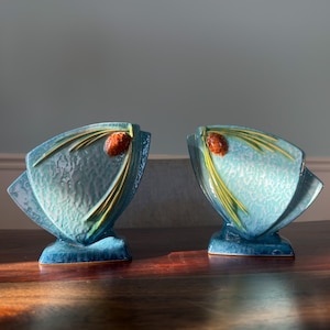 May include: Two matching teal-colored ceramic vases with a textured surface. Each vase features a stylized design of a pine cone and needles in shades of orange and green. The vases are displayed on a dark wooden surface.